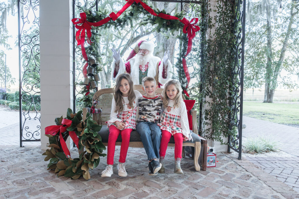 Three siblings sit on a bench smiling, while not knowing that Santa is standing behind them.