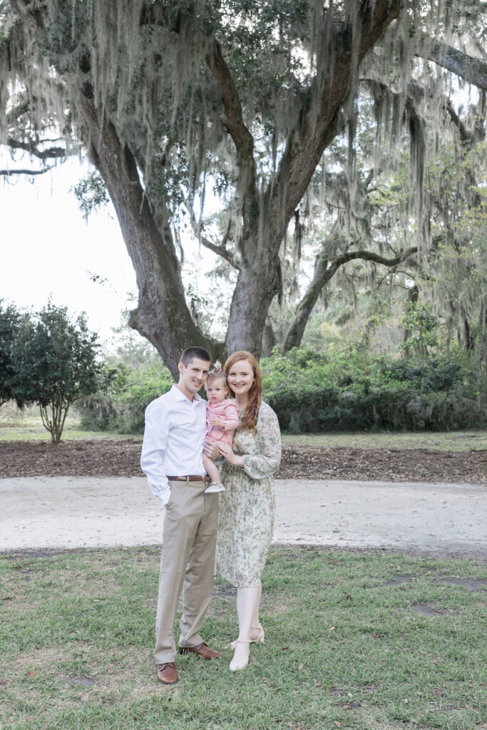 A family of three smile in front of the tree where the mom and dad were married at HollyOaks on the Marsh.