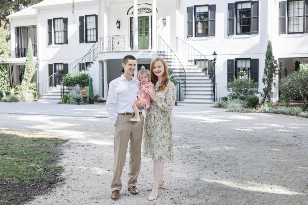 A young couple with their toddler daughter take fall family portraits in front of the manor house at HollyOaks on the Marsh.