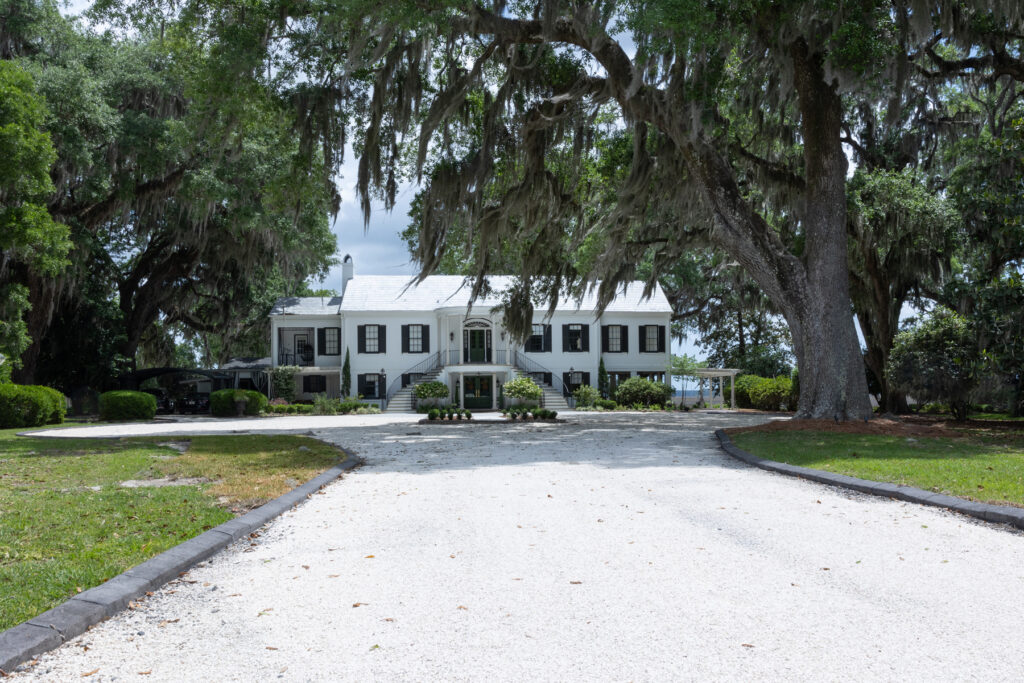 A front view of the long, gravel drive, oak trees, and white manor house at HollyOaks on the Marsh.