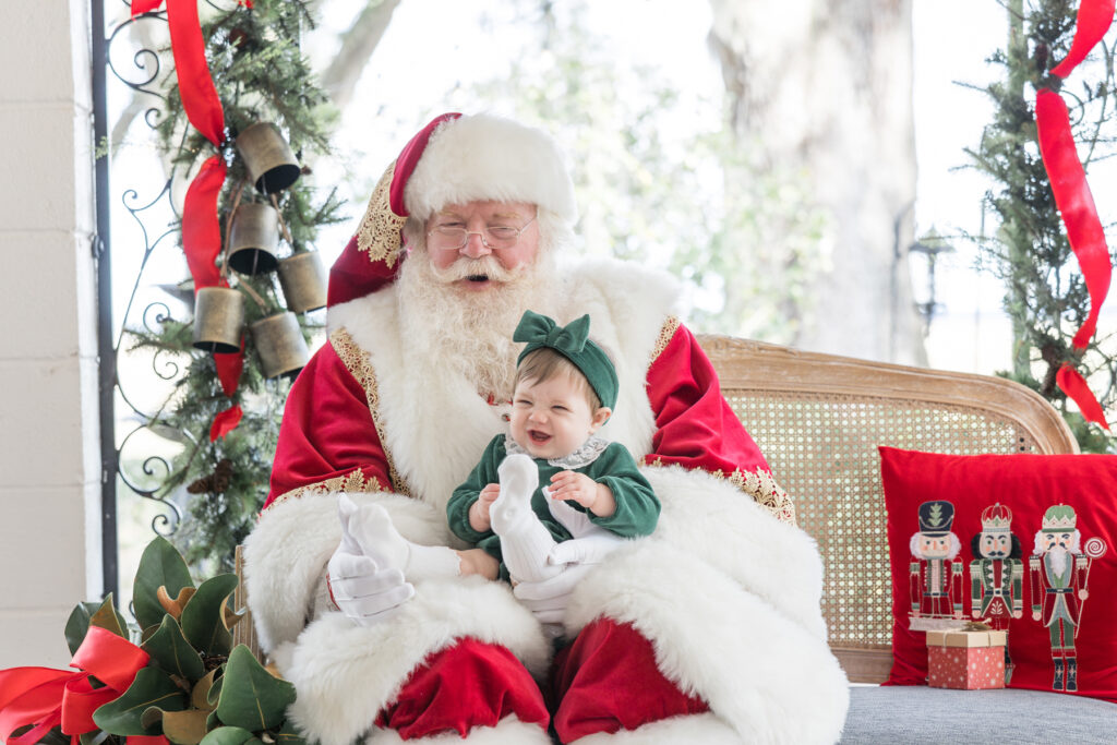 Santa and a baby girl smile sitting on a bench in Christmas portrait setup area at HollyOaks on the Marsh.