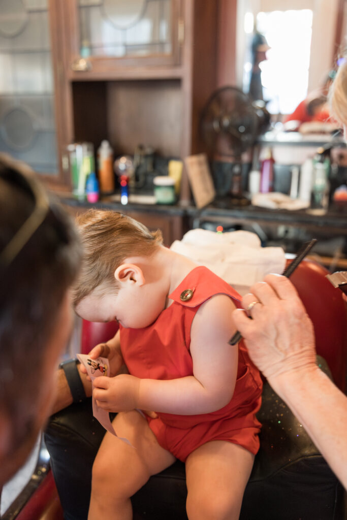 A toddler boy puts his head down as he is sitting in a salon chair. 