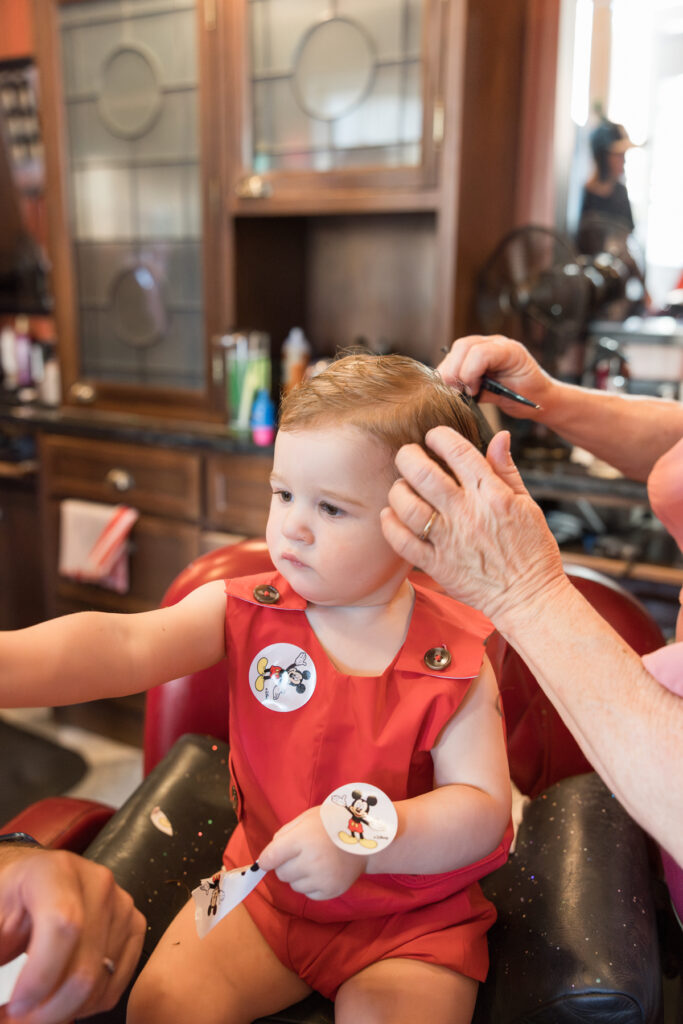 A toddler, dressed in a red jon jon from Littles on Liberty in Savannah, has his haircut while he puts Mickey stickers all over him.