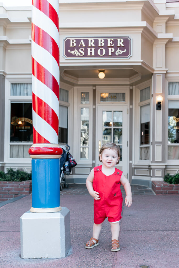 A toddler boy dressed in a Jack Keene Richmond Red jon jon from The Beaufort Bonnet Company smiles in front of the Harmony Barbershop before he has his haircut.