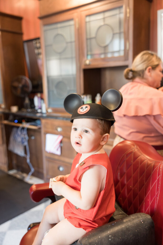 A little boy sits in a salon chair with a Mickey Mouse hat on.