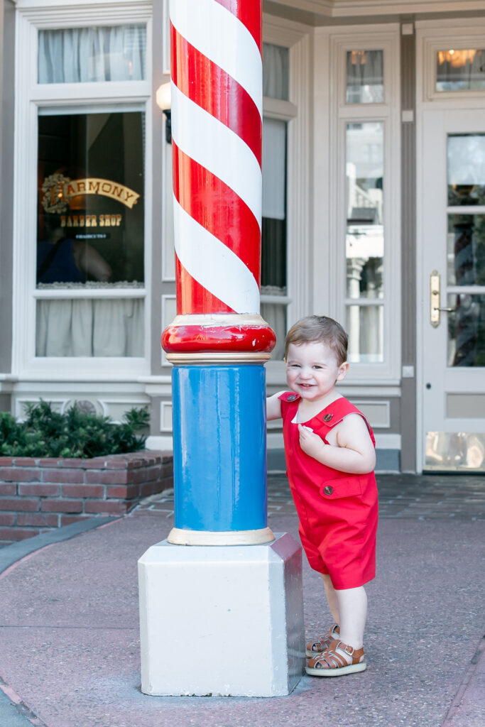 Toddler boy dressed in red jon jon from Littles on Liberty standing by barbershop pole at Disney's Magic Kingdom, smiling at his Savannah Family Photographer mom.