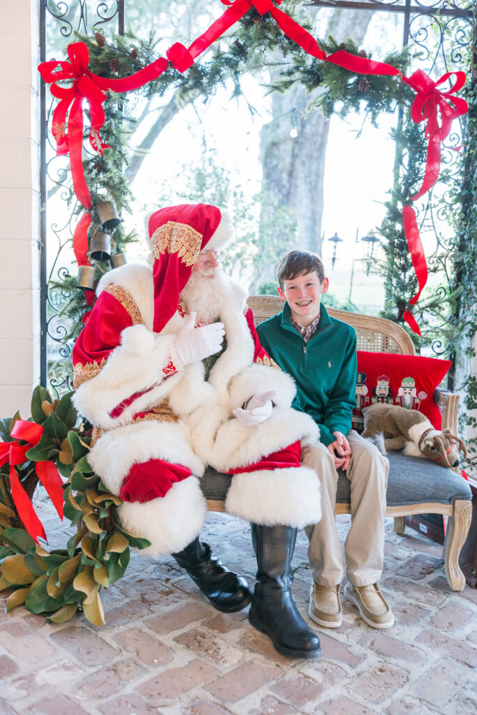 A pre-teen boy in a green pullover smiles as Santa, dressed in traditional red, talks to him.