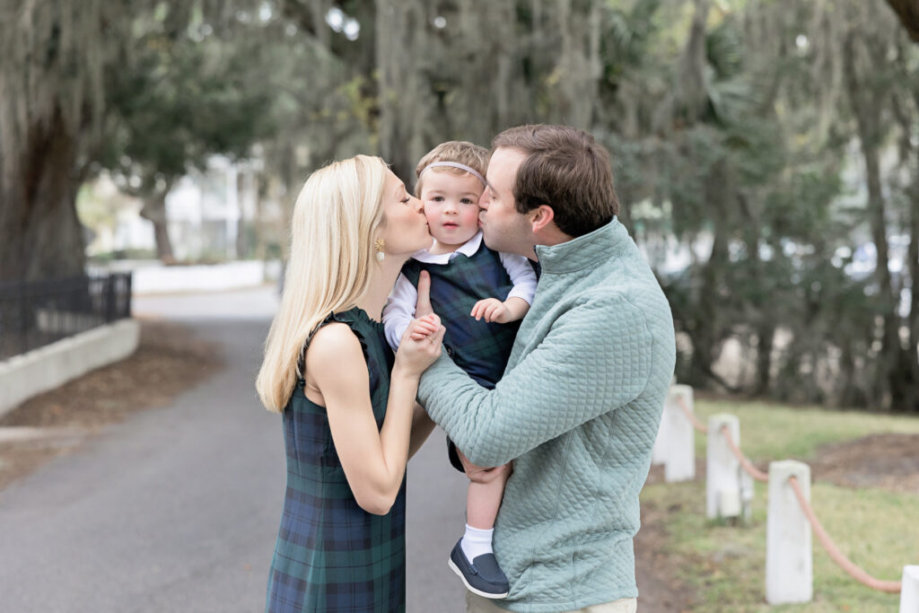 A little boy is given a kiss by his parents during a family photography session during fall in Savannah, Georgia at Bluff Drive on Isle of Hope.