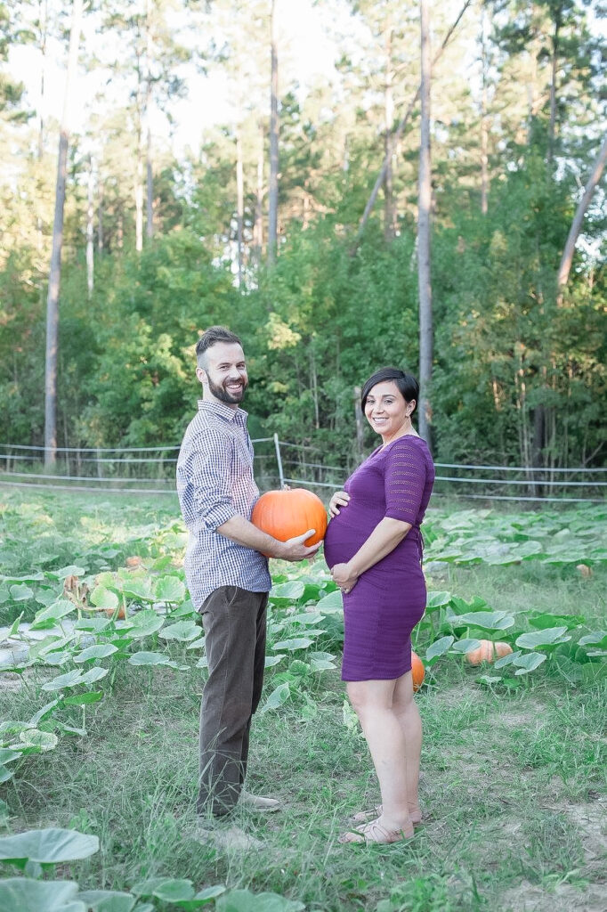 Expectant parents pose with a pumpkin in between in fall at a pumpkin patch.