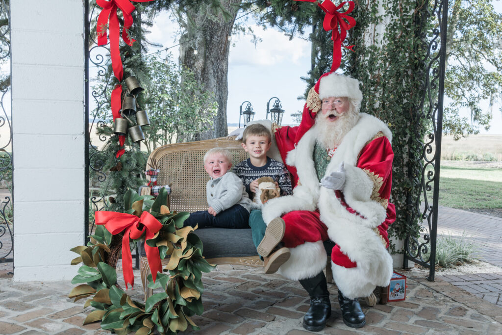 Two boys and Santa sit on a bench on a veranda with marsh views. The littlest one is screaming and the oldest is smiling.