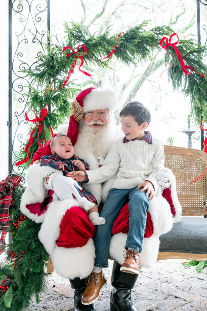 A baby cries sitting in Santa's lap while his brother consoles him.