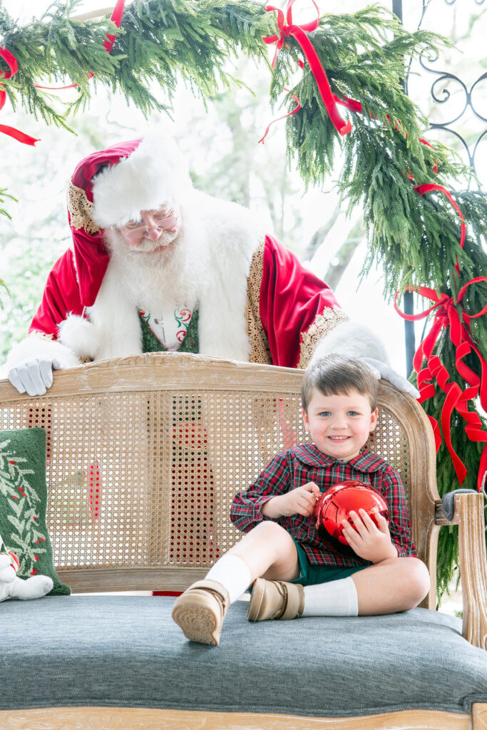 A young boy holds a red Christmas ornament while Santa stands behind him looking on. 