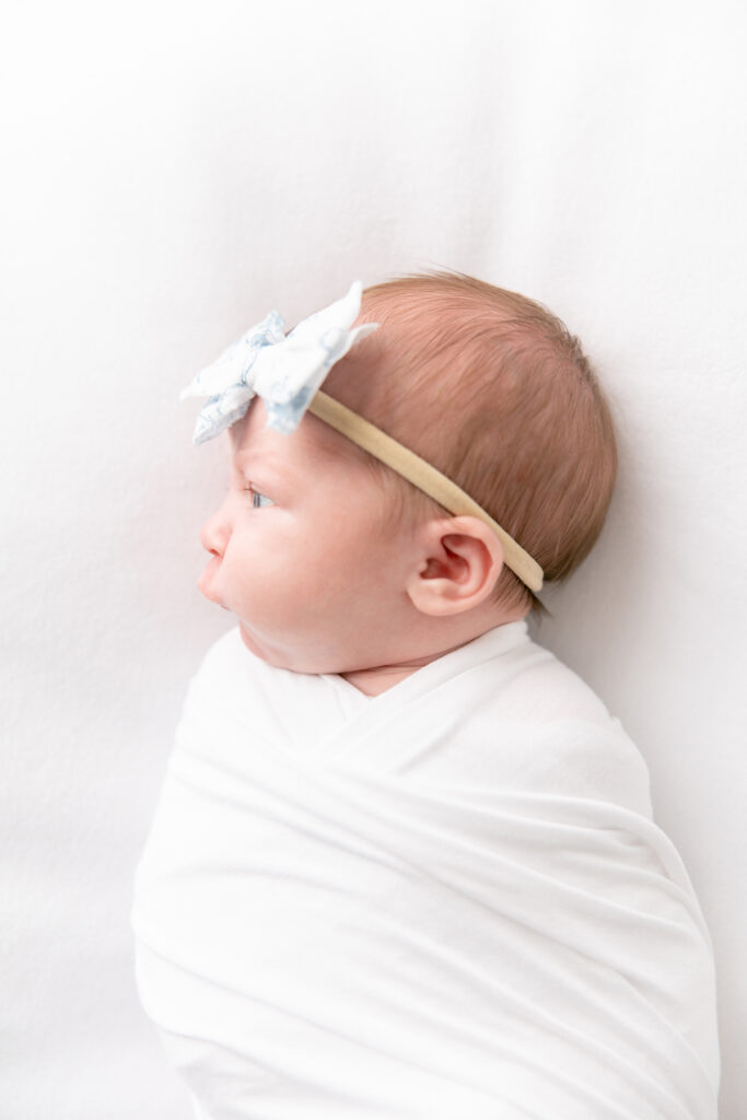 Side profile picture of a newborn girl with a blue and white bow on a white background swaddled in a white wrap during a classic newborn studio session with Savannah maternity photographer, Tiffany Bradley.