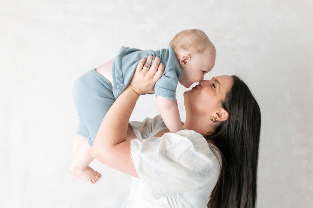 A mom makes a kissy face while holding her little boy in the air close to her face. 
