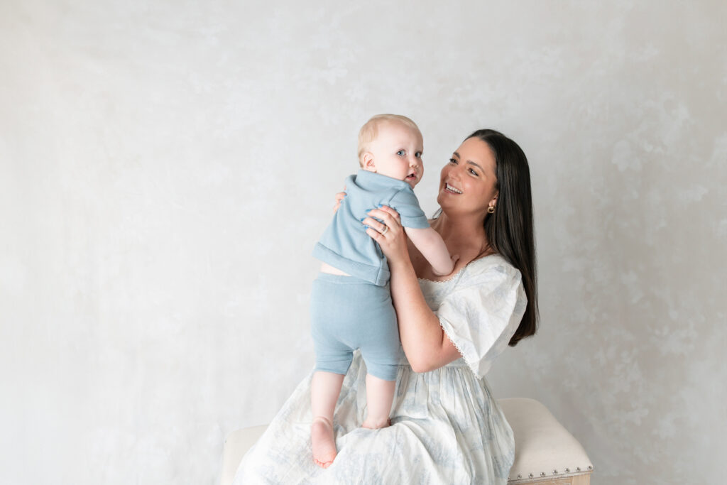 A baby boy looks back at the camera as him mom holds him up and smiles at him. 