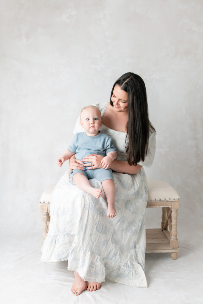 During a mommy and me photoshoot, a mom looks down at her baby boy while sitting on a tan bench. 