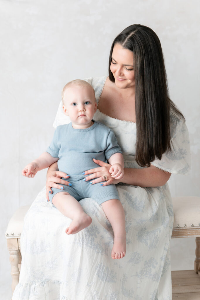 A one-year old baby boy with blue eyes and blonde hair stares into the camera while sitting on his mom's lap.