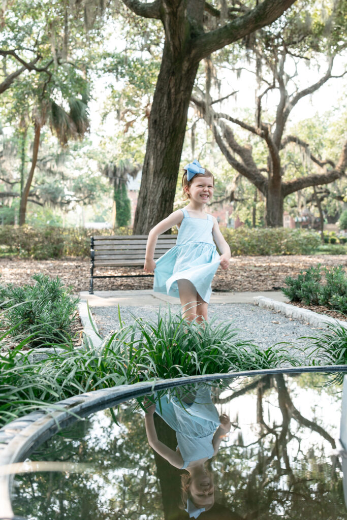 A little girl in a blue dress spins in front of a fountain in Ardsley Park in Savannah, Georgia during a summer photo session. 