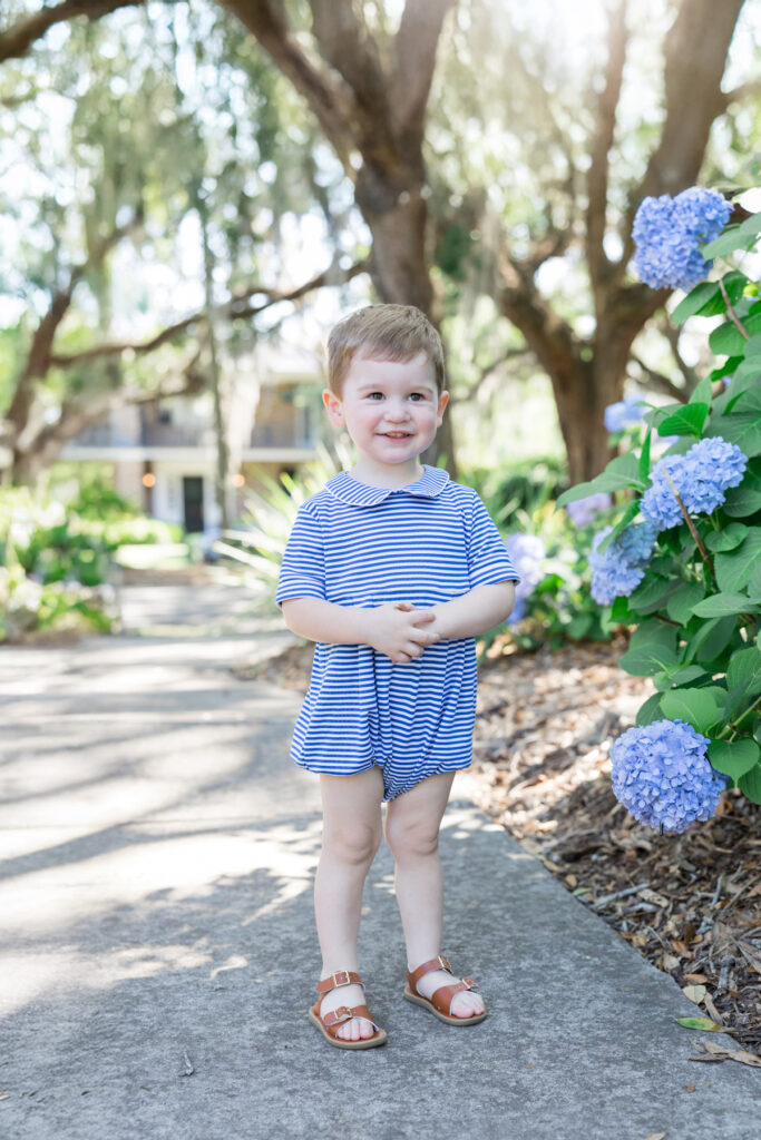 A two-year-old boy in a blue and white striped Beaufort Bonnet bubble outfit stands next to beautiful blue hydrangeas in Savannah, Georgia during summertime.