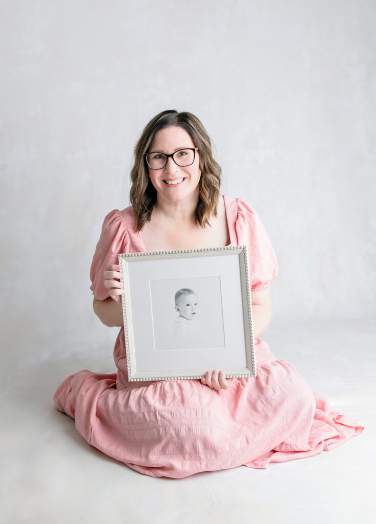 Tiffany Bradley, a Savannah newborn photographer smiles holding a framed portrait from a child's heirloom portrait session.