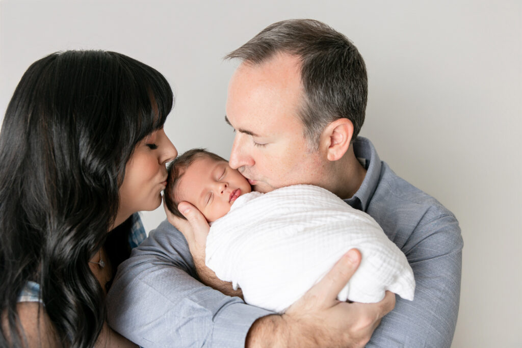 A new mom and dad kiss their baby during a studio portrat session in Savannah, GA with photographer Tiffany Bradley.