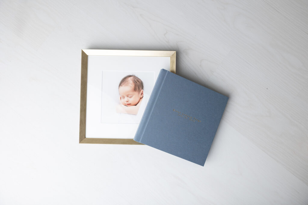A blue linen album and a gold framed print of a newborn from a portrait session with Tiffany Bradley Photography in Savannah.