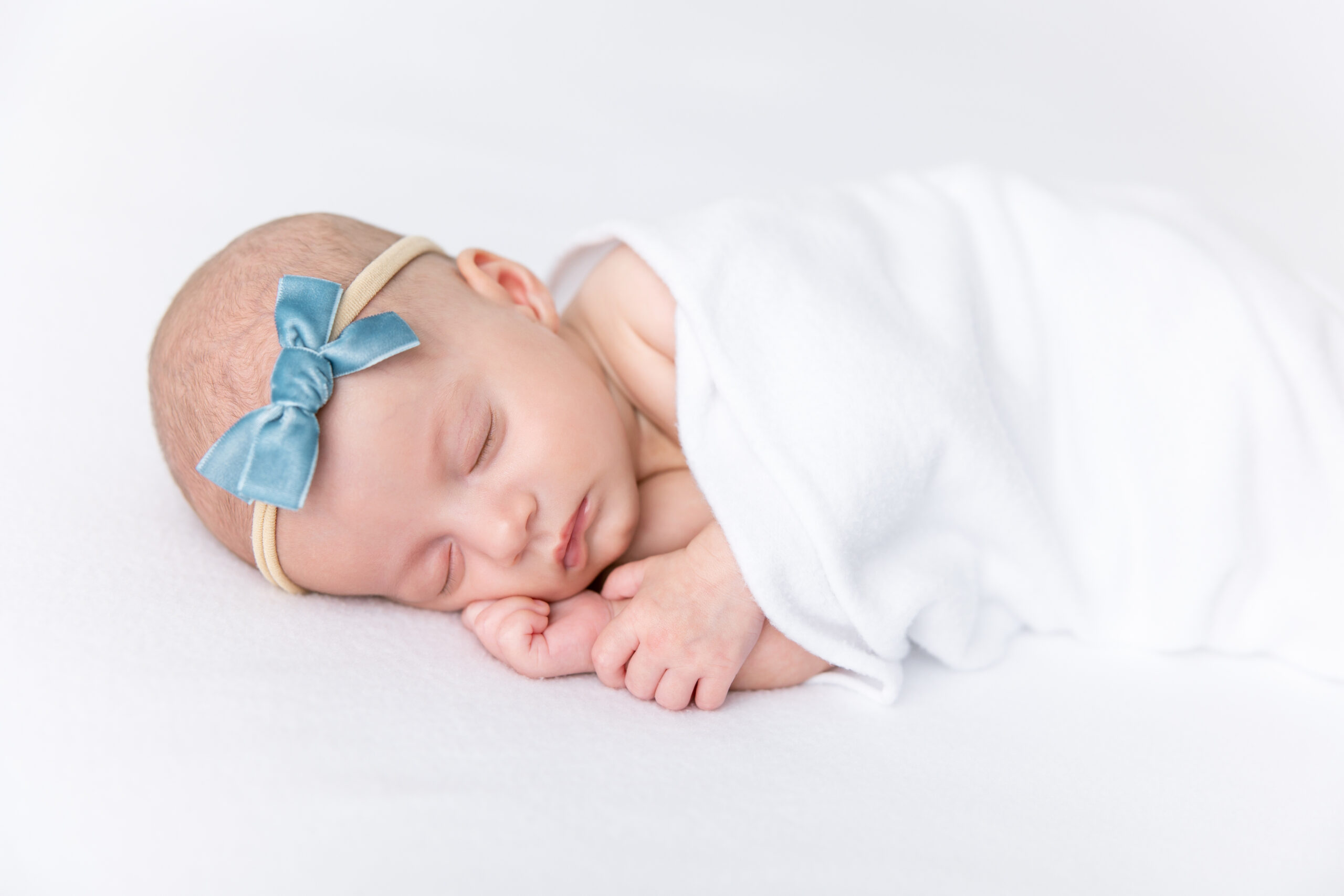 A newborn sleeps quietly snuggled in a cozy white blanket with a blue silk bow on her head, showcasing newborn photo safety in her pose.