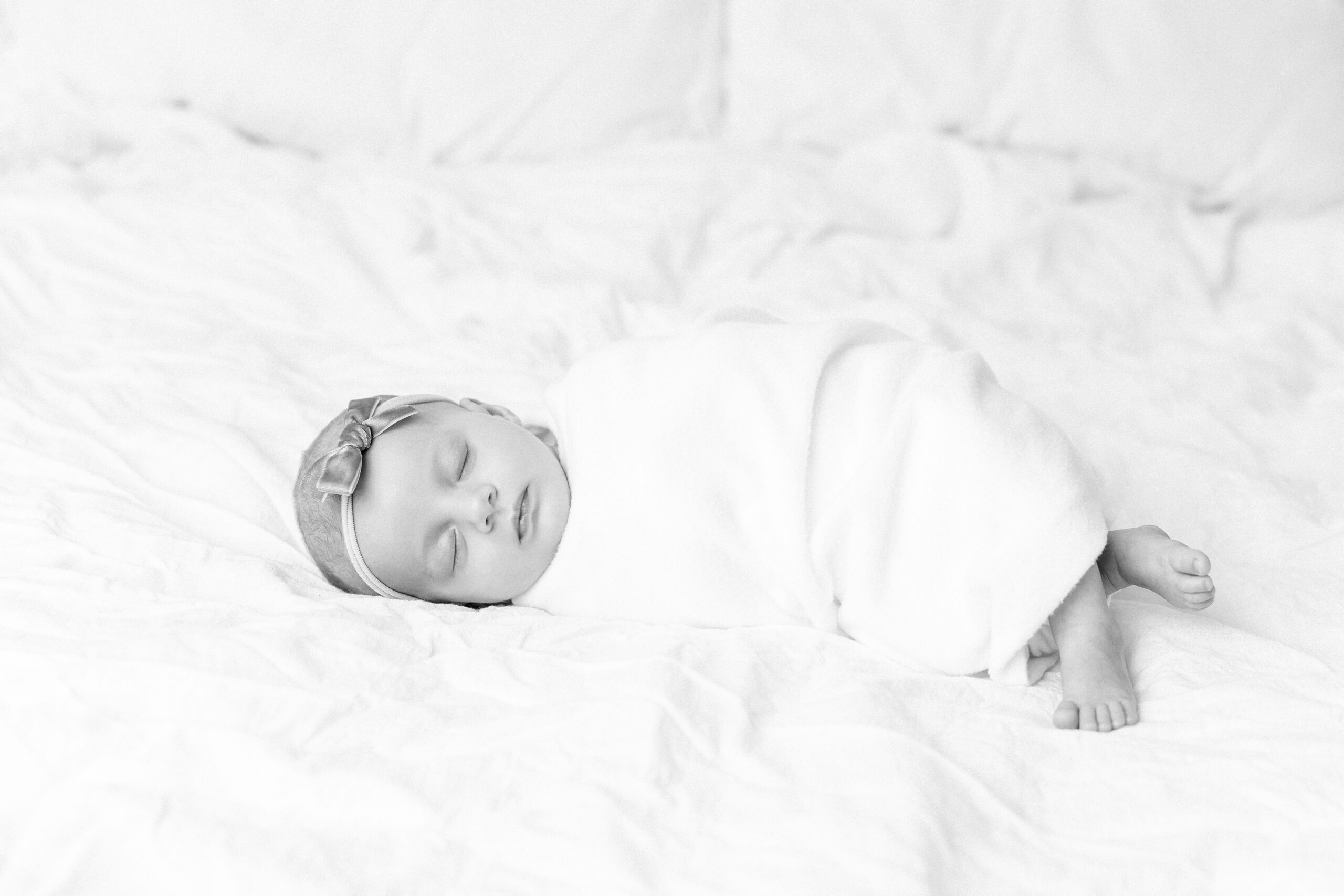 A black and white image of a newborn with feet peeping out from a cozy white blanket with posing that follows newborn photography safety guidelines.