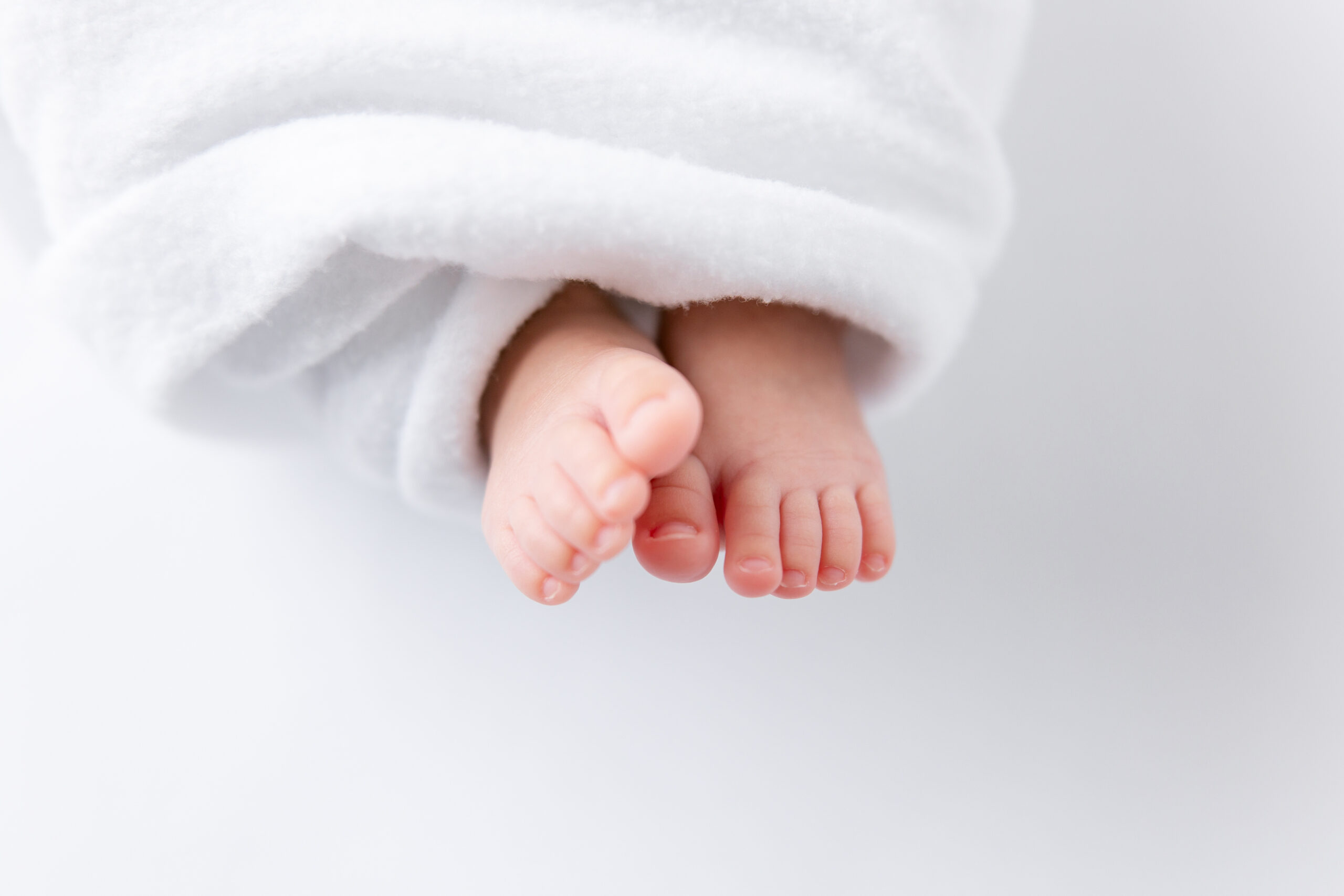 Sweet little baby feet pop out from under a cozy white blanket while prioritizing newborn posing safety.