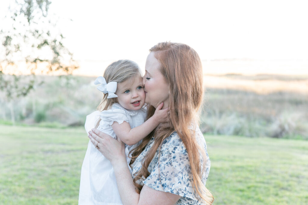 Mom kisses her daughter in front of a marsh view at HollyOaks on the Marsh during a Savannah family photography session with Tiffany Bradley