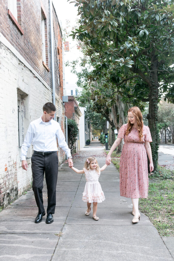 A family of 3 walk down a side walk toward Chatham Square in Savannah, GA for a maternity portrait session with Tiffany Bradley Photography.