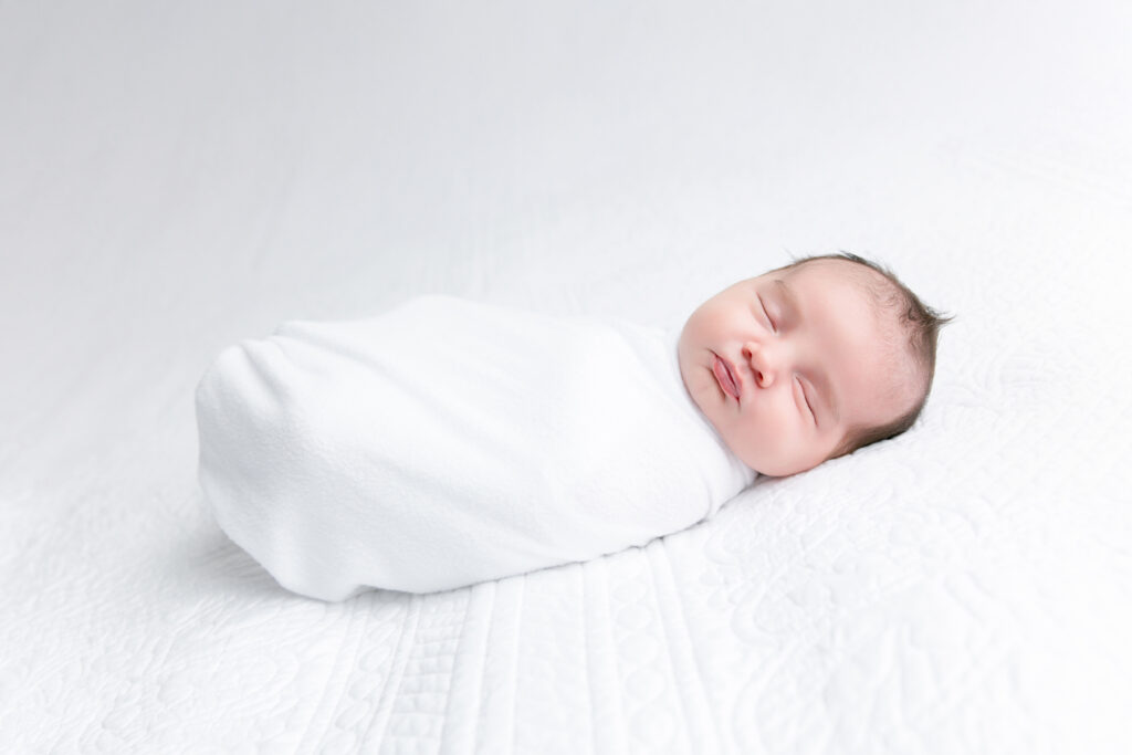 Newborn baby boy wrapped in white on a quilted white blanket during a newborn photography session in Savannah, GA.