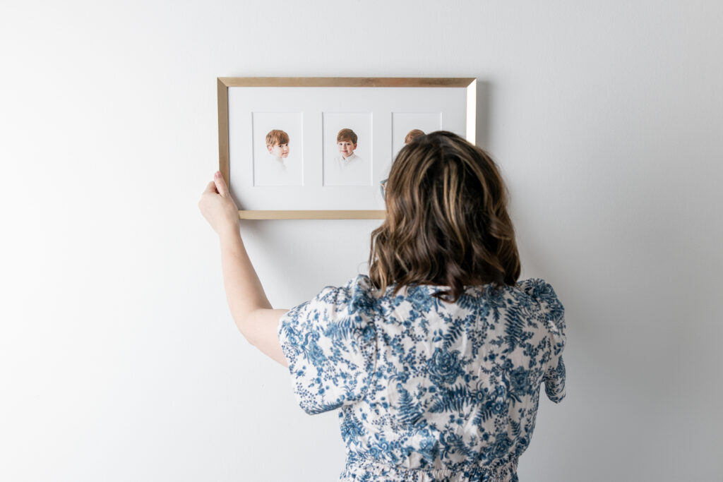 Tiffany Bradley, heirloom portrait photographer, hangs a trio panel frame of heirloom children's portraits on a white wall.