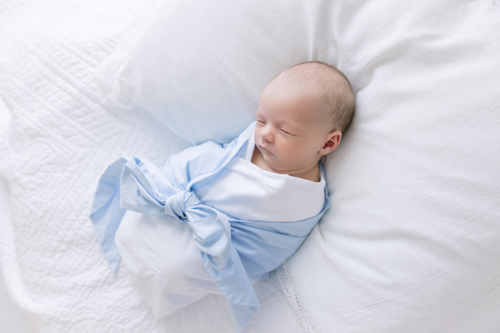 Heirloom newborn portrait of baby in blue bow swaddle from Littles on Liberty on a white pillow and heirloom blanket.