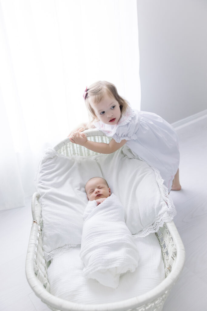 Baby in Pottery Barn bassinet in front of a window while toddler sister climbs on bassinet. Savannah GA Newborn Photography
