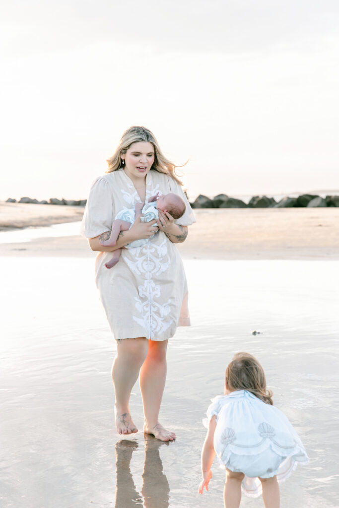 Mom walking along beach with her newborn son and toddler daughter.