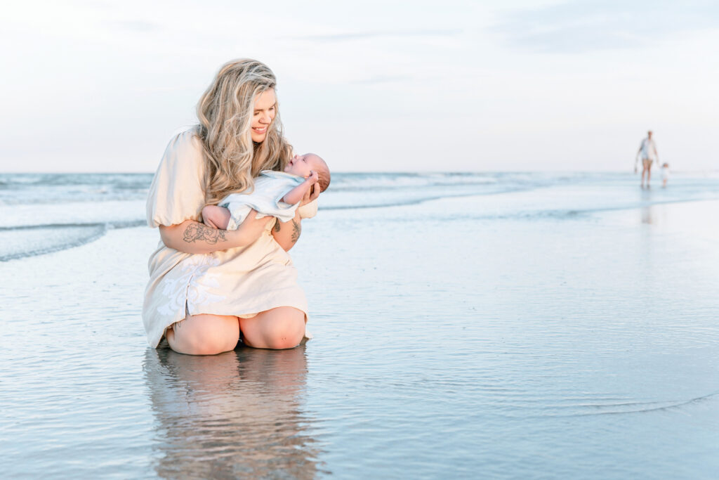 Mom and newborn sitting by ocean while dad and daughter walk in the distance during Tybee island photography session with Tiffany Bradley Photography